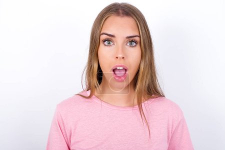 Photo for Emotional attractive Young Caucasian girl wearing pink T-shirt on white background with opened mouth expresses great surprise and fright, stares at camera. Unexpected shocking news and human reaction. - Royalty Free Image