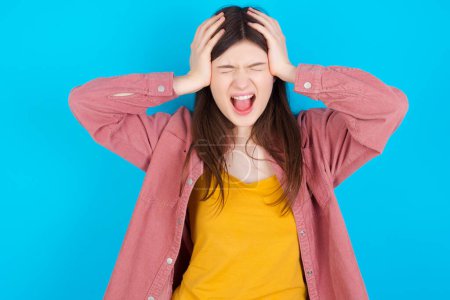 Photo for Shocked panic young caucasian girl wearing pink shirt isolated over blue background holding hands on head and screaming in despair and frustration. - Royalty Free Image