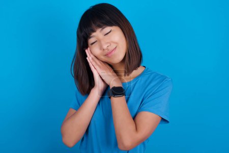 Téléchargez les photos : Jeune asiatique femme portant bleu t-shirt sur fond bleu se penche sur pressé paumes ferme les yeux et a agréable sourire rêves sur quelque chose - en image libre de droit