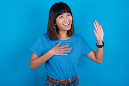 Téléchargez les photos : Promets-moi de ne pas regretter. Portrait de sincère jeune femme asiatique portant un t-shirt bleu sur fond bleu levant un bras et tenant la main sur le cœur comme donner serment, dire la vérité, vous voulez croire. - en image libre de droit