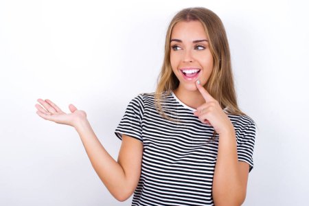 Photo for Positive beautiful blonde girl wearing striped t-shirt on white background advert promo touch finger teeth - Royalty Free Image