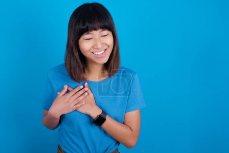 Téléchargez les photos : Jeune femme asiatique portant bleu t-shirt sur fond bleu exprime le bonheur, rit agréablement, garde les mains sur le cœur - en image libre de droit