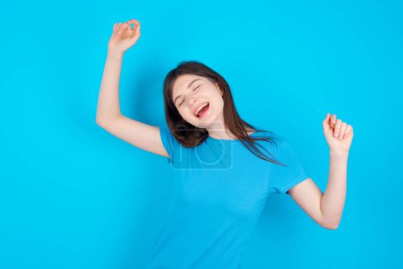 Photo for Photo of upbeat young caucasian girl wearing blue t-shirt isolated over blue studio background has fun and dances carefree wear being in perfect mood makes movements. Spends free time on disco party - Royalty Free Image
