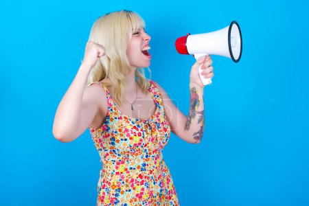 Photo for Caucasian girl wearing floral dress isolated over blue background communicates shouting loud holding a megaphone, expressing success and positive concept, idea for marketing or sales. - Royalty Free Image