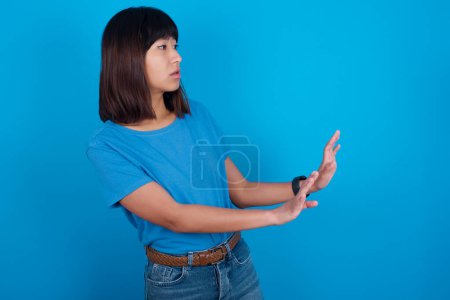 Foto de Disgustado joven asiático mujer usando azul camiseta contra azul fondo mantiene las manos hacia vacío espacio y pide no acercarse ve algo desagradable - Imagen libre de derechos
