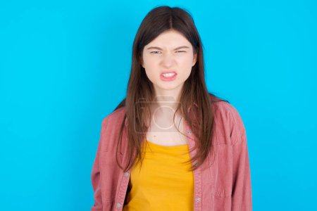 Photo for Mad crazy young caucasian girl wearing pink shirt isolated over blue background clenches teeth angrily, being annoyed with coming noise. Negative feeling concept. - Royalty Free Image