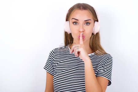 Photo for Beautiful blonde girl wearing striped t-shirt on white background making hush gesture with finger on her lips wearing  wireless headphones. Be quiet. - Royalty Free Image