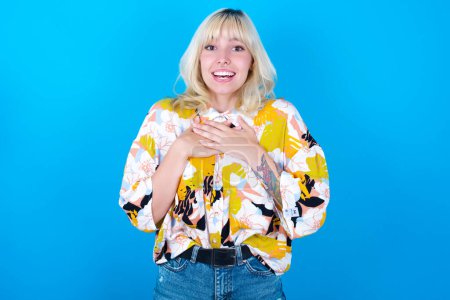 Photo for Happy smiling caucasian girl wearing floral shirt isolated over blue background has hands on chest near heart. Human emotions, real feelings and facial expression concept. - Royalty Free Image