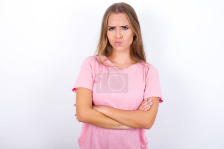 Foto de Imagen de una joven caucásica enojada con una camiseta rosa sobre fondo blanco cruzando brazos. Mirando a la cámara con expresión decepcionada. - Imagen libre de derechos