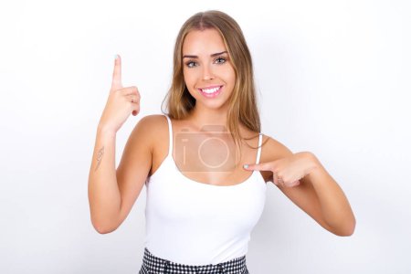 Photo for Young Caucasian girl wearing white tank top on white background says: wow how exciting it is, has amazed expression, indicates something. One hand on her chest and pointing with other hand. - Royalty Free Image