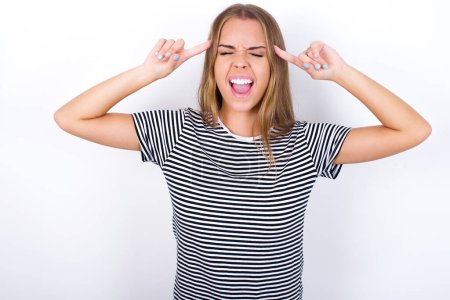 Photo for Photo of crazy beautiful blonde girl wearing striped t-shirt on white background screaming and pointing with fingers at hair closed eyes - Royalty Free Image