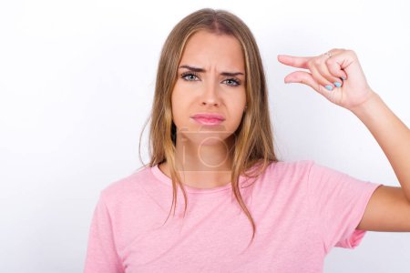 Photo for Upset Young Caucasian girl wearing pink T-shirt on white background shapes little gesture with hand demonstrates something very tiny small size. Not very much - Royalty Free Image
