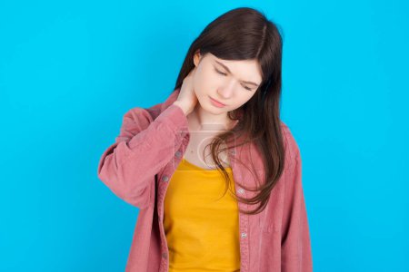 Foto de Joven chica caucásica con camisa rosa aislada sobre fondo azul que sufre de lesión dolor de espalda y cuello, tocando el cuello con la mano, dolor muscular. - Imagen libre de derechos