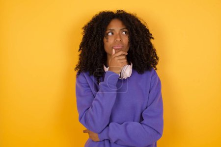 Téléchargez les photos : Portrait de jeune femme afro-américaine réfléchie avec écouteurs sur fond jaune - en image libre de droit