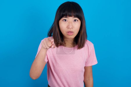 Foto de Sorprendido joven mujer asiática con camiseta rosa contra los puntos de fondo azul frente con el dedo índice en la cámara y. Concepto de sorpresa y publicidad. - Imagen libre de derechos