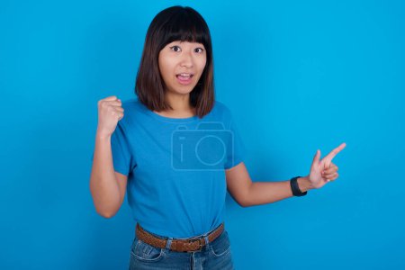 Foto de Joven asiático mujer usando azul camiseta contra azul fondo puntos en vacío espacio sosteniendo puño arriba, ganador gesto. - Imagen libre de derechos