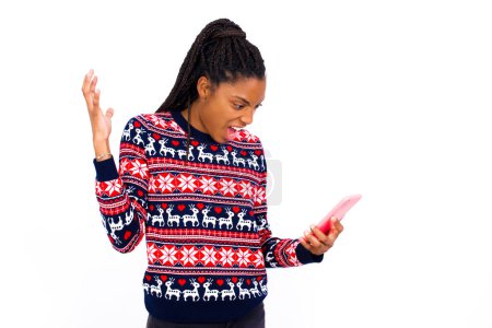 Téléchargez les photos : Photo de femme afro-américaine indignée et indignée portant un pull de Noël contre un mur blanc tient un téléphone portable, appelle, discute avec un collègue, exprime des émotions négatives. Les gens et la colère. - en image libre de droit