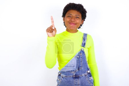 Photo for Young African American woman with short hair wearing denim overall against white wall showing and pointing up with fingers number one while smiling confident and happy. - Royalty Free Image