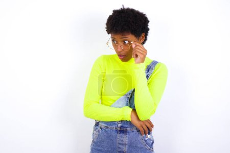 Foto de Mujer afroamericana joven asombrada con el pelo corto usando denim en general contra la pared blanca se ve a un lado sorprendentemente con la boca abierta. - Imagen libre de derechos
