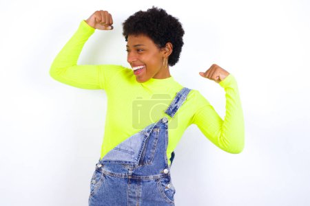 Téléchargez les photos : Oui, je suis gagnant. Portrait de charmante jeune femme afro-américaine ravie et excitée avec les cheveux courts portant le denim dans l'ensemble contre le mur blanc levant le poing dans le triomphe et la victoire souriant atteindre le succès en souriant de plaisir. - en image libre de droit