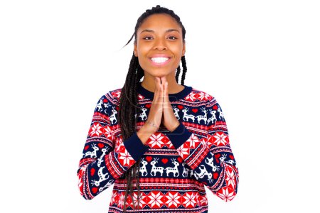 Photo for African American woman wearing Christmas sweater against white wall keeps palms together, has pleased expression. Glad attractive male makes request, pleads for mercy. Hopeful young adult. - Royalty Free Image