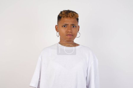 Photo for Pensive attractive young African American woman wearing white t-shirt being deep in thoughts, raises eyebrows, curves lips, wears fashionable clothes, stands against gray wall with copy space for your text. - Royalty Free Image