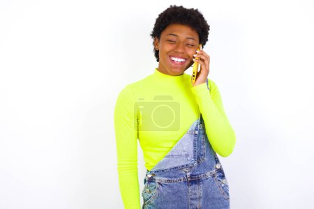 Foto de Divertida joven afroamericana con el pelo corto vistiendo denim general contra la pared blanca ríe felizmente, tiene conversación telefónica, ser divertido por amigo, cierra los ojos. - Imagen libre de derechos