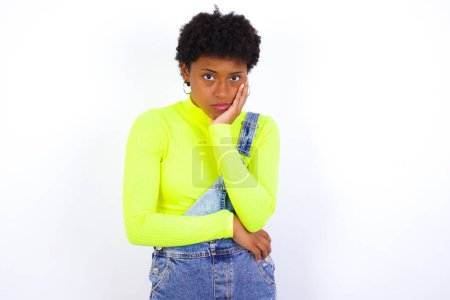 Photo for Very bored young African American woman with short hair wearing denim overall against white wall holding hand on cheek while support it with another crossed hand, looking tired and sick. - Royalty Free Image