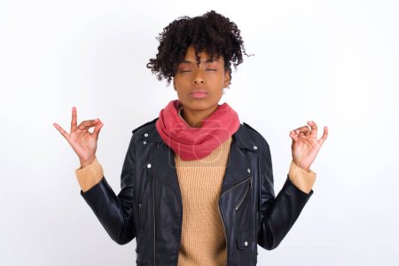 Young beautiful African American woman wearing biker jacket against white wall doing yoga, keeping eyes closed, holding fingers in mudra gesture. Meditation, religion and spiritual practices.
