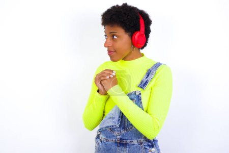 Foto de Joven mujer afroamericana con el pelo corto con denim en general contra la pared blanca lleva auriculares estéreo escuchando música concentrada y mirando a un lado con interés. - Imagen libre de derechos