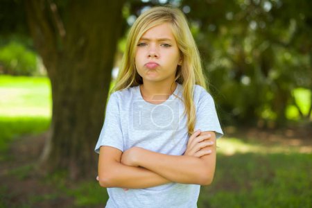 Gloomy dissatisfied caucasian little kid girl wearing white t-shirt standing outdoor in the park looks with miserable expression at camera from under forehead, makes unhappy grimace