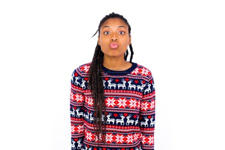 Téléchargez les photos : Femme afro-américaine portant un pull de Noël contre un mur blanc garde les lèvres comme allant embrasser quelqu'un, a une expression heureuse, visage grimaçant. Debout dedans. Concept de beauté. - en image libre de droit
