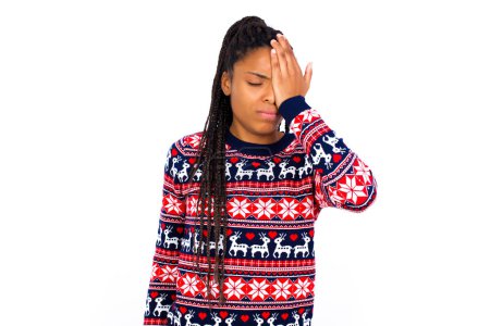 Foto de Mujer afroamericana frustrada vistiendo suéter de Navidad contra pared blanca sosteniendo la mano en la frente deprimida lamentando lo que hizo teniendo dolor de cabeza, luciendo estresante. - Imagen libre de derechos