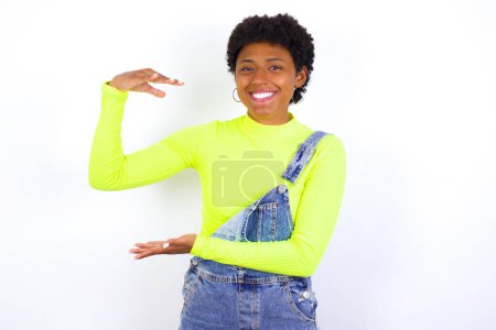Foto de Joven mujer afroamericana con el pelo corto con denim en general contra la pared blanca gesto con las manos mostrando signo de tamaño grande y grande, símbolo de medida. - Imagen libre de derechos