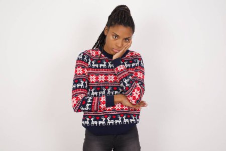 Foto de Mujer afroamericana muy aburrida usando suéter de Navidad contra pared blanca agarrada de la mano en la mejilla mientras la apoya con otra mano cruzada, luciendo cansada y enferma. - Imagen libre de derechos