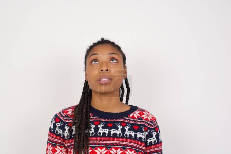 Foto de Mujer afroamericana vistiendo suéter de Navidad contra la pared blanca mirando hacia arriba mientras ve algo extraño. - Imagen libre de derechos