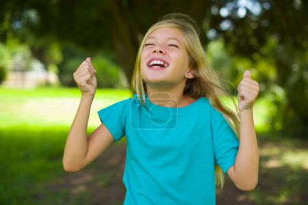 Porträt eines schönen kaukasischen kleinen Mädchens mit blauem T-Shirt, das im Park steht und sich glücklich fühlt