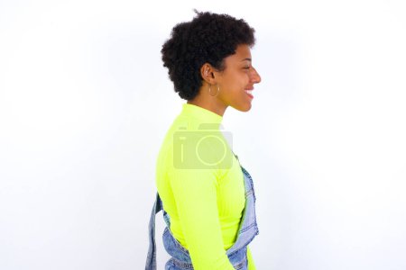 Photo for Profile of smiling young African American woman with short hair wearing denim overall against white wall with healthy skin, has contemplative expression, ready to have outdoor walk. - Royalty Free Image