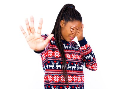 Foto de Mujer afroamericana vistiendo suéter de Navidad contra la pared blanca cubre los ojos con la palma y haciendo gesto de parada, trata de esconderse. No me mires, no quiero ver, me da vergüenza o miedo. - Imagen libre de derechos