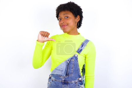 Foto de Primer plano de la joven mujer afroamericana alegre con el pelo corto con denim en general contra la pared blanca se ve alegre, satisfecho y confiado, apunta a sí mismo con el pulgar. - Imagen libre de derechos