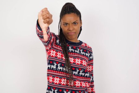 Téléchargez les photos : Mécontentement femme afro-américaine portant un pull de Noël contre un mur blanc montre signe de désapprobation, maintient le pouce vers le bas, exprime son aversion, fronce les sourcils visage dans le mécontentement. Sentiments négatifs. - en image libre de droit