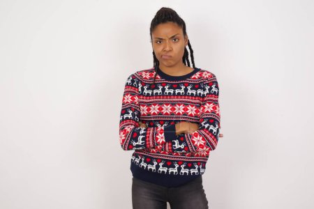 Téléchargez les photos : Photo de femme afro-américaine en colère portant un pull de Noël contre un mur blanc croisant les bras. Regardant la caméra avec une expression déçue. - en image libre de droit