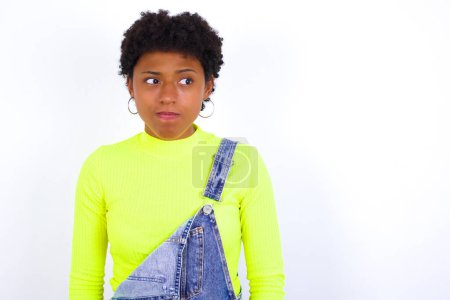 Photo for Amazed puzzled young African American woman with short hair wearing denim overall against white wall curves lips and has worried look, sees something awful in front. - Royalty Free Image