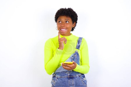 Foto de Imagen de una joven afroamericana pensante soñando con el pelo corto usando denim en general contra la pared blanca usando el teléfono móvil y sosteniendo la mano en la cara. Toma de decisiones y concepto de redes sociales. - Imagen libre de derechos