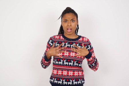 Téléchargez les photos : Femme afro-américaine portant un pull de Noël contre un mur blanc garde les mains sur la poitrine choquée et effrayée, la bouche largement ouverte, regarde la caméra en disant : Qui, moi? - en image libre de droit