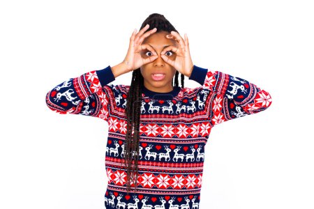 Foto de Juguetona mujer afroamericana emocionada vistiendo suéter de Navidad contra pared blanca mostrando signo Ok con ambas manos en los ojos, fingiendo usar gafas. - Imagen libre de derechos