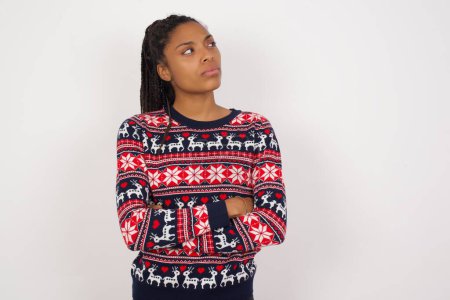 Téléchargez les photos : Image de femme afro-américaine bouleversée portant un pull de Noël contre un mur blanc avec les bras croisés. Regarder avec expression déçue de côté après avoir écouté les mauvaises nouvelles. - en image libre de droit