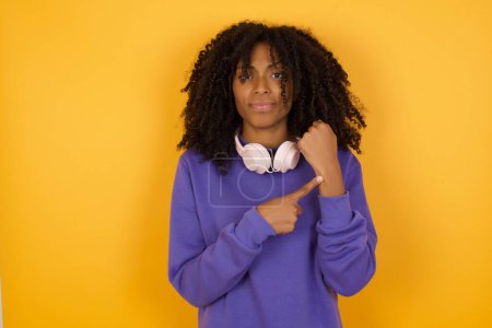 Photo for Portrait of young expressive african american woman with headphones on yellow background pointing at wrist, time management concept - Royalty Free Image