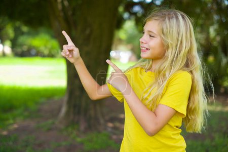 Téléchargez les photos : Caucasien petite fille portant jaune t-shirt debout à l'extérieur sourire excité doigts directifs regarder vide espace - en image libre de droit