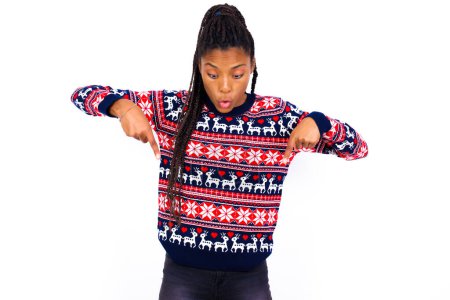 Foto de La mujer afroamericana asombrada que usa suéter de Navidad contra la pared blanca apunta hacia abajo con los dedos delanteros, abre la boca sorprendida. Concepto de publicidad. - Imagen libre de derechos